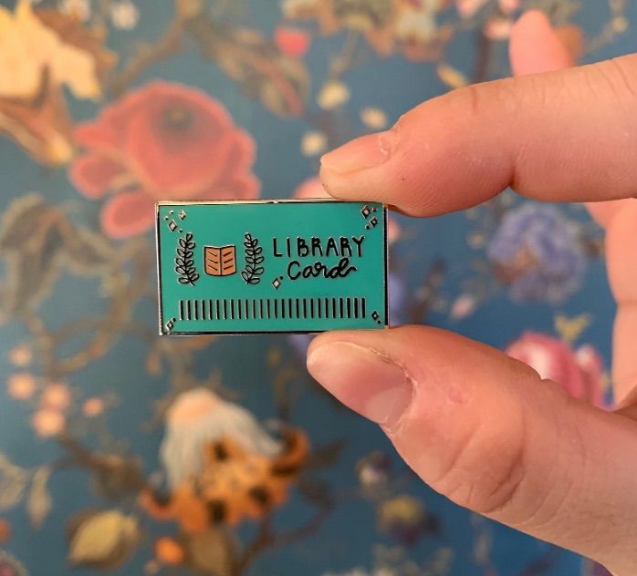 Image of a green enamel pin held by white fingers. It says "library card" and looks like a library card. 