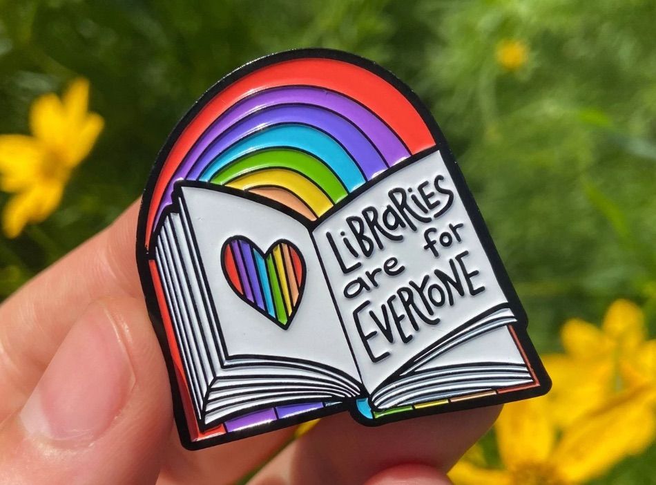 Image of a pin that is an open book. It has a rainbow above it, and it says "libraries are for everyone."