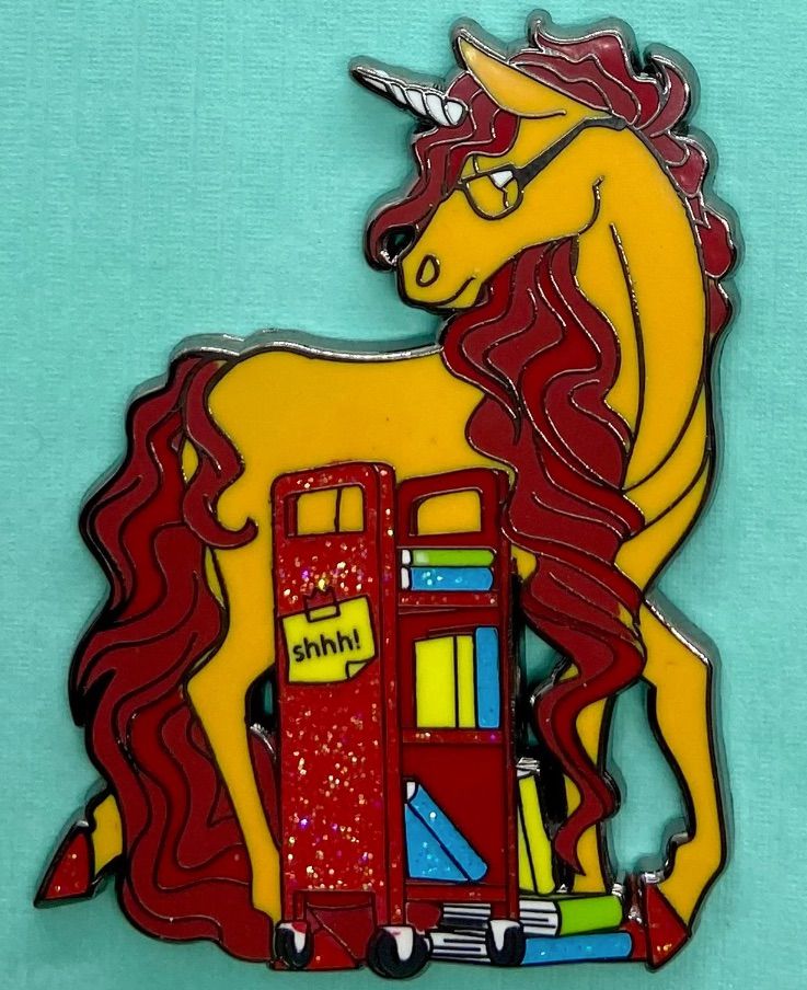 Image of an enamel pin that features a colorful unicorn and a book cart