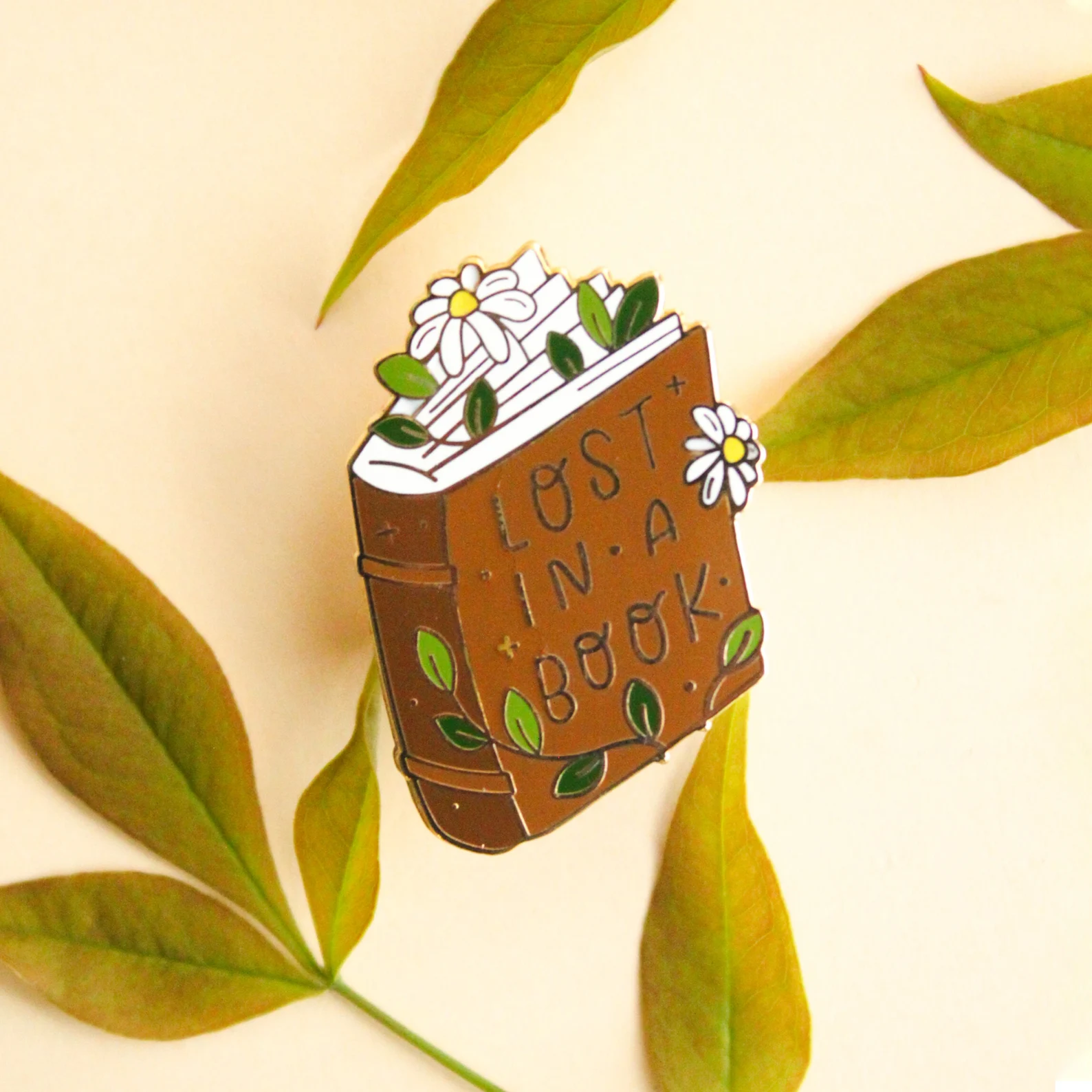 An enamel pin of a book decorate in daisies that reads "Lost in a book"