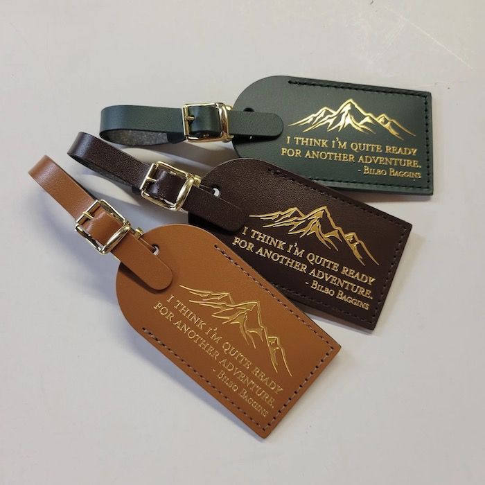 three luggage tags in gold, brown, and green leather. Each tag contains a Lord of the Rings quote in gold