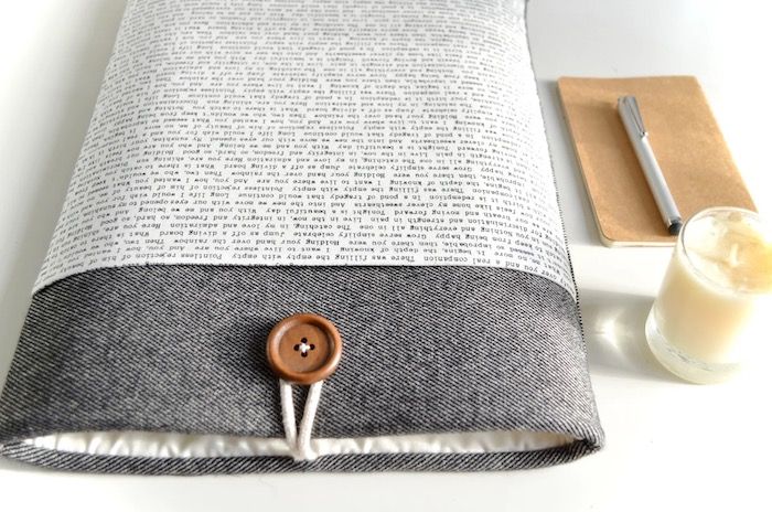 a charcoal grey book sleeve with a button closure and a pocket in a word pattern. the sleeve is next to a notice candle and a small pen and notebook
