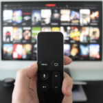 a hand holding a remote with a TV on a streaming service menu out of focus in the background