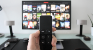 a photo of someone holding a remote with a TV on a streaming service menu out of focus in the background