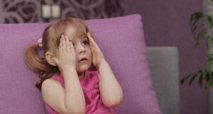 一个坐在紫色沙发上看电视的小孩，双手举在脸上，表情惊恐