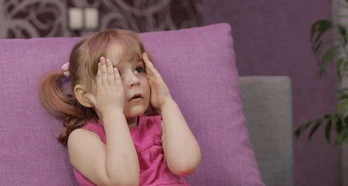 a small child sitting on a purple couch while watching TV, hands held up to their face in a scared expression