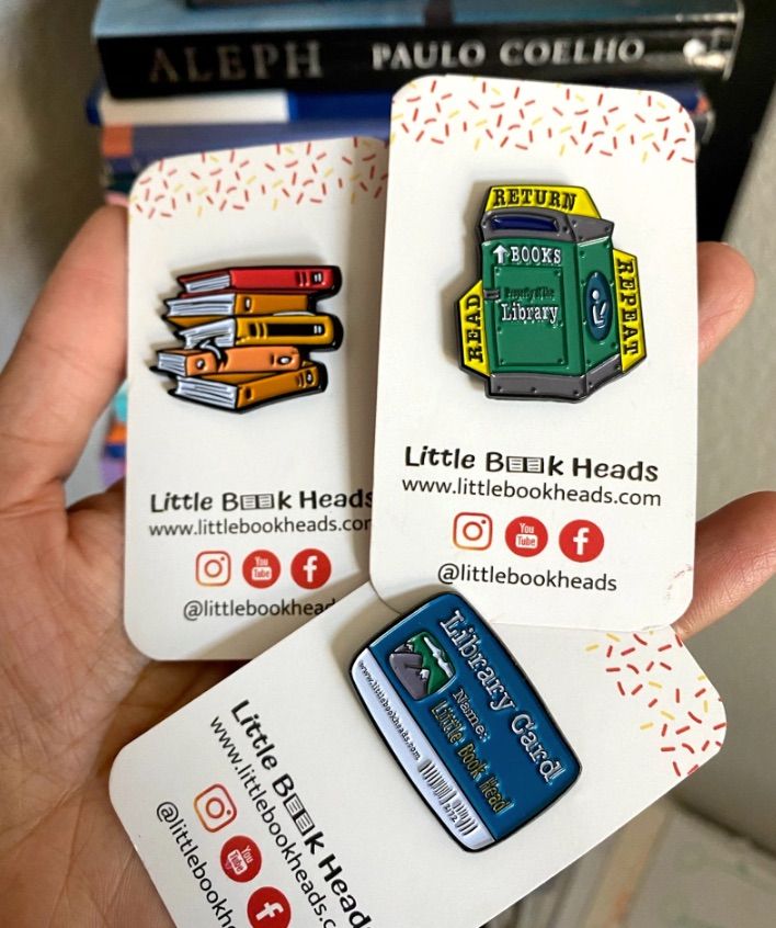 Set of three enamel pins, including a book stack, a library book return, and a library card. 