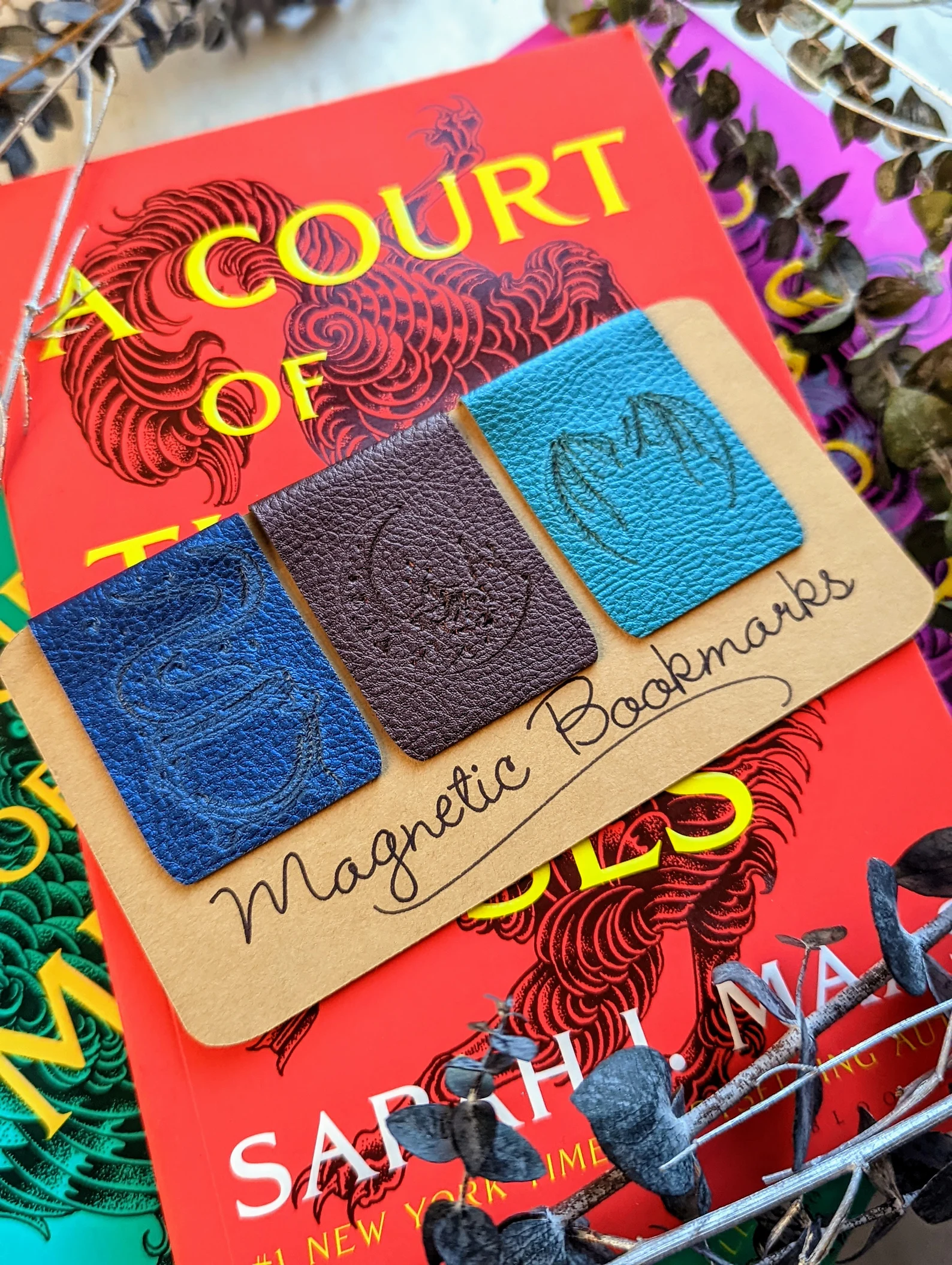 A photo of 3 leather magnetic bookmarks on a piece of paper that reads magnetic bookmarks. Left to right, the bookmarks are blue with a cauldren, brown with a moon, and teal with bat wings. The bookmarks are staged on top of the book A court of throns and roses.