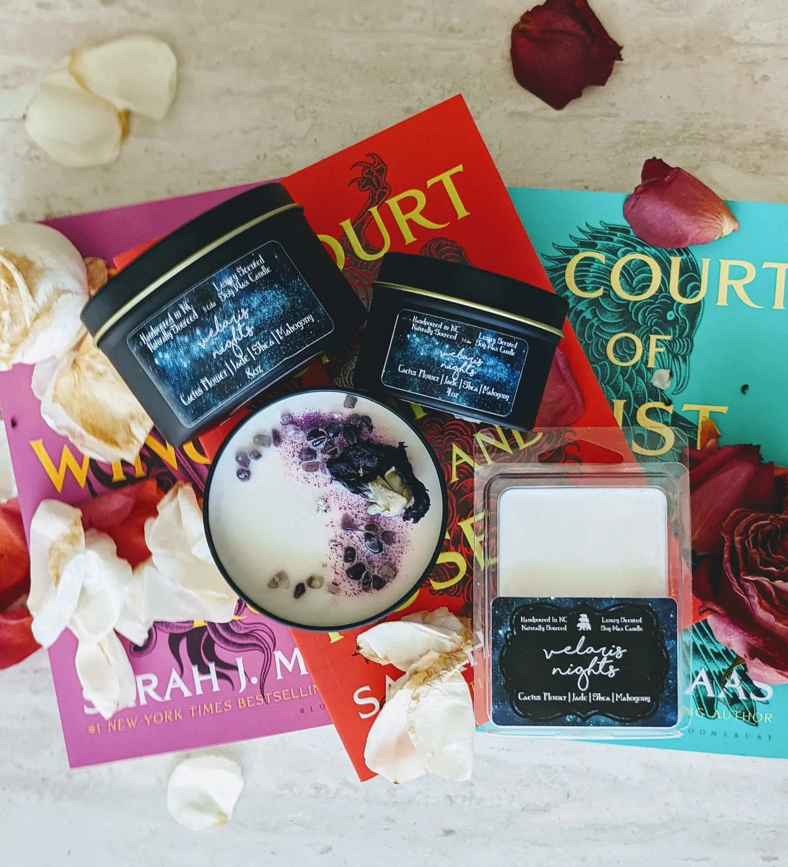 A photo of three candles in black metal containers and a wax block that read Velaris nights cactus flowers, jade, shea, and mahogany. The candles are staged on top of the three books in the court of thorns and roses series and rose petals.