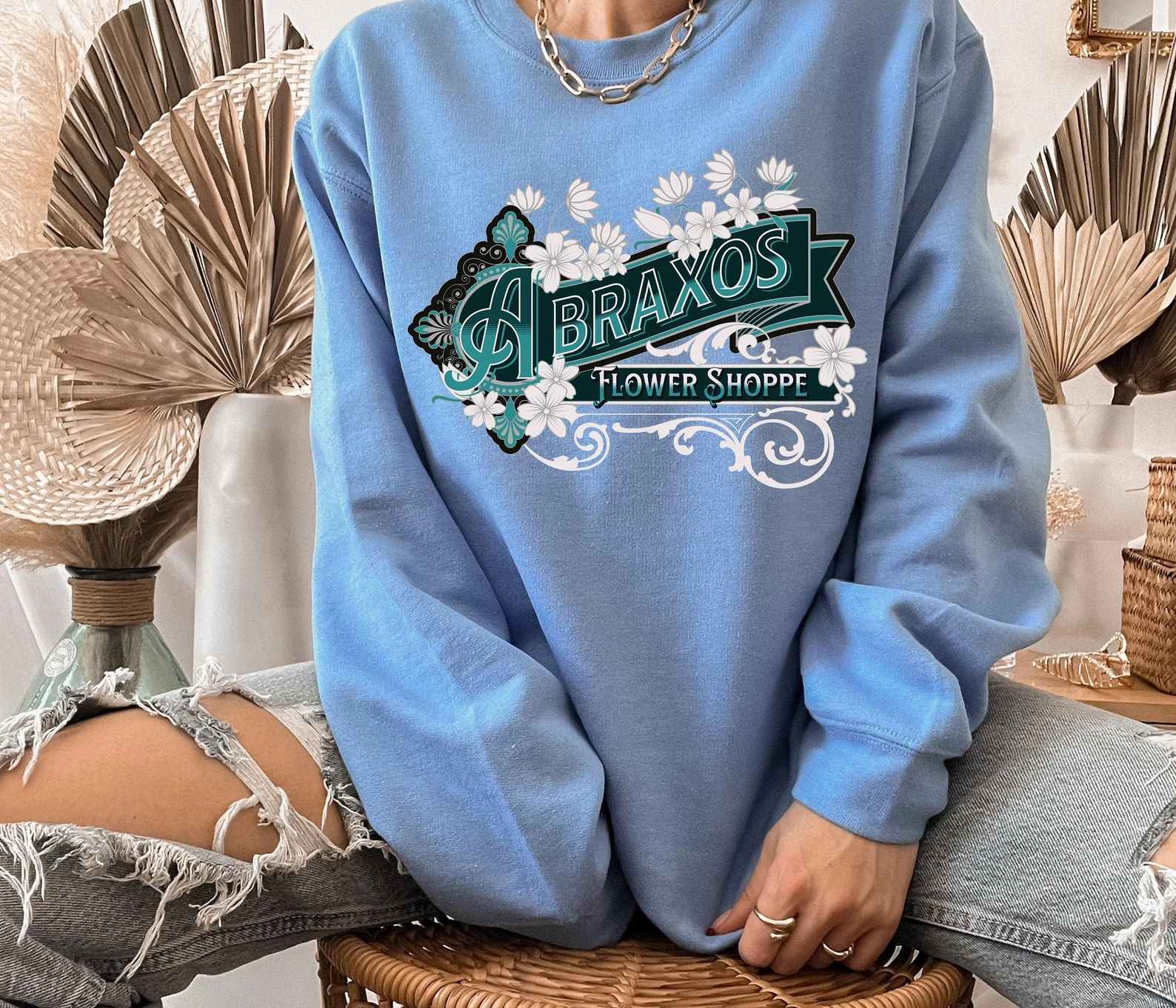 A photo of a person sitting on a stool wearing a blue sweatshirt with the words Abraxos Flower Shoppe written on it. 