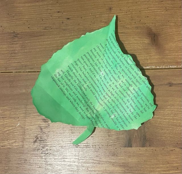 A leaf cut out of an old book page has been painted green and is sitting on a wooden table.