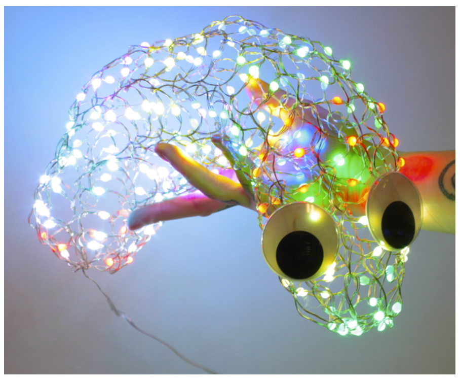 A knitted blob of LED lights, with googly eyes, is held in the hand of its creator. 