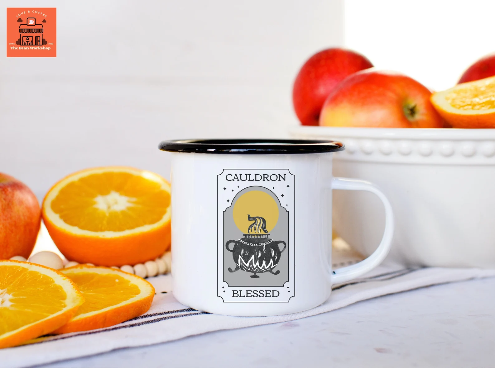 A photo of a white mug with a black rim that says Cauldron Blessed on the front with a yellow moon, a black cauldron, and gray accents. Left to right, the mug is staged with oranges on the table and apples in a bowl.