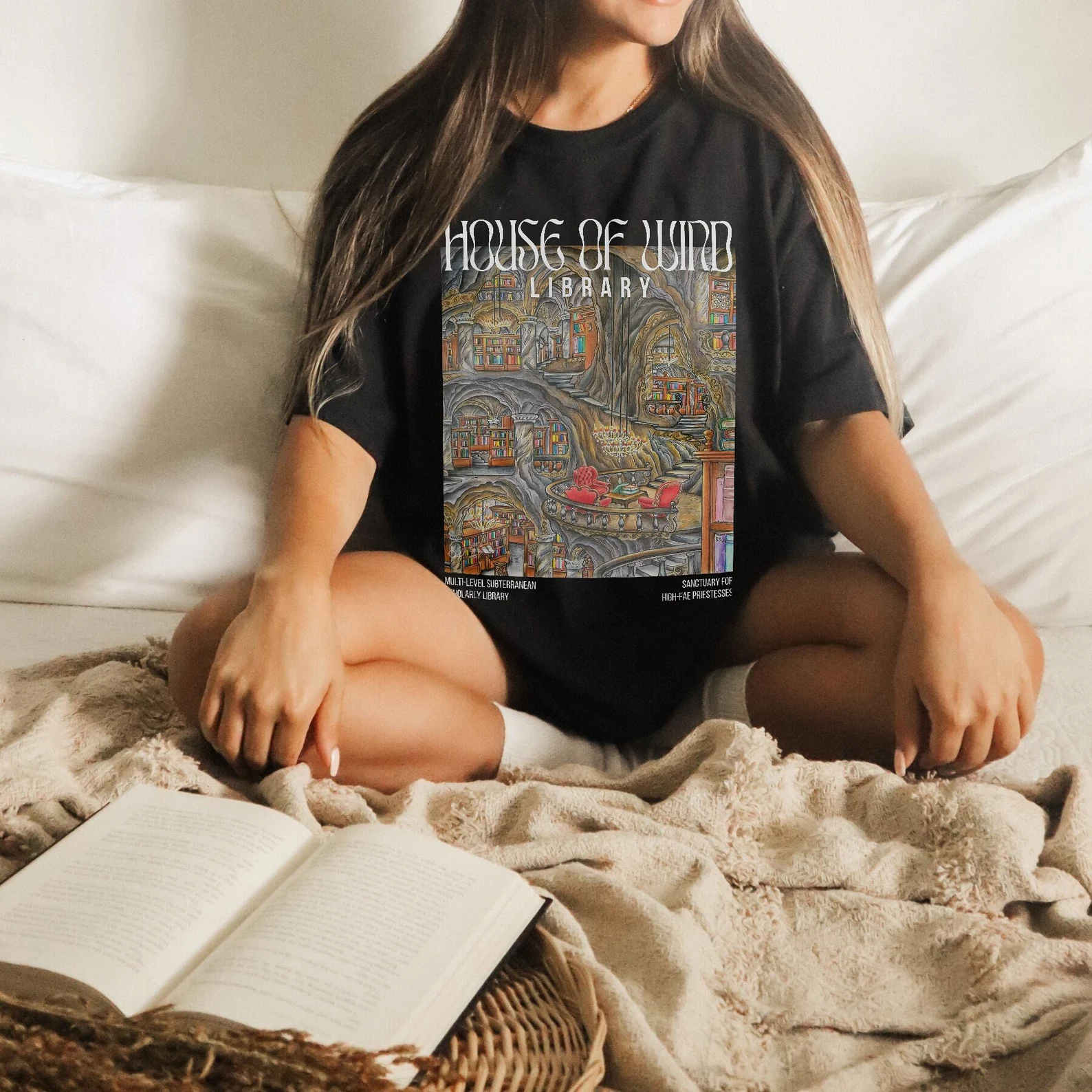 A photo of a person with long brown hair sitting on a bed wearing a black shirt with a rectangular image of a library and the words House Of Wind Library at the top and Multi-level subterranean Scholarly library and Sanctuary for High-fae priestesses at the bottom. 