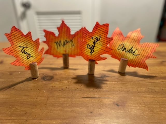 Colorful painted leaves from old book pages standing upright on corks, each with a name written in black, on a wooden table