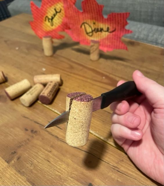A small knife is cutting halfway through a wine cork. More corks and colorful leaves are in the background.