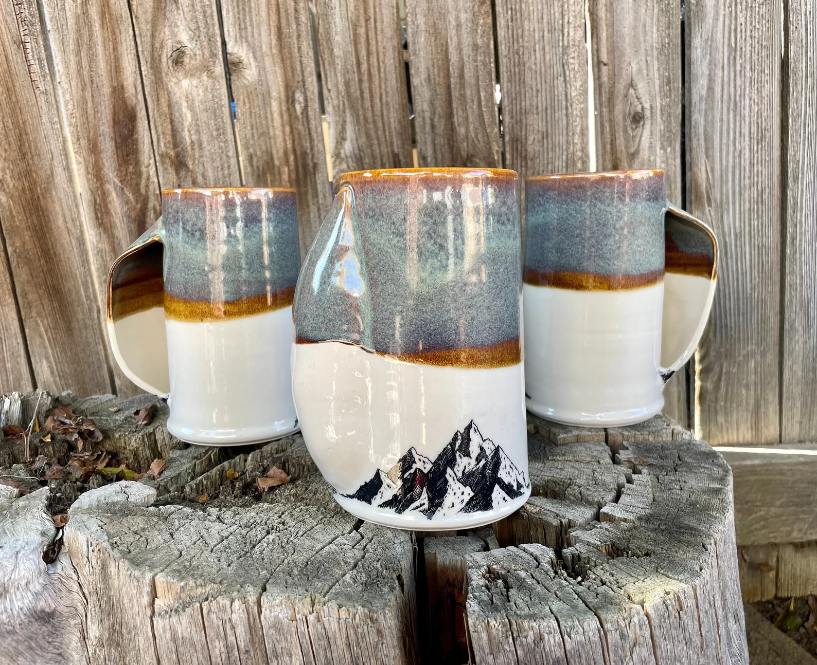 A photo of three mugs with a pocket handle with a blue top half and a white bottom half. The image of black mountains is on the bottom half of the mug. The mugs are staged on a wooden stump.