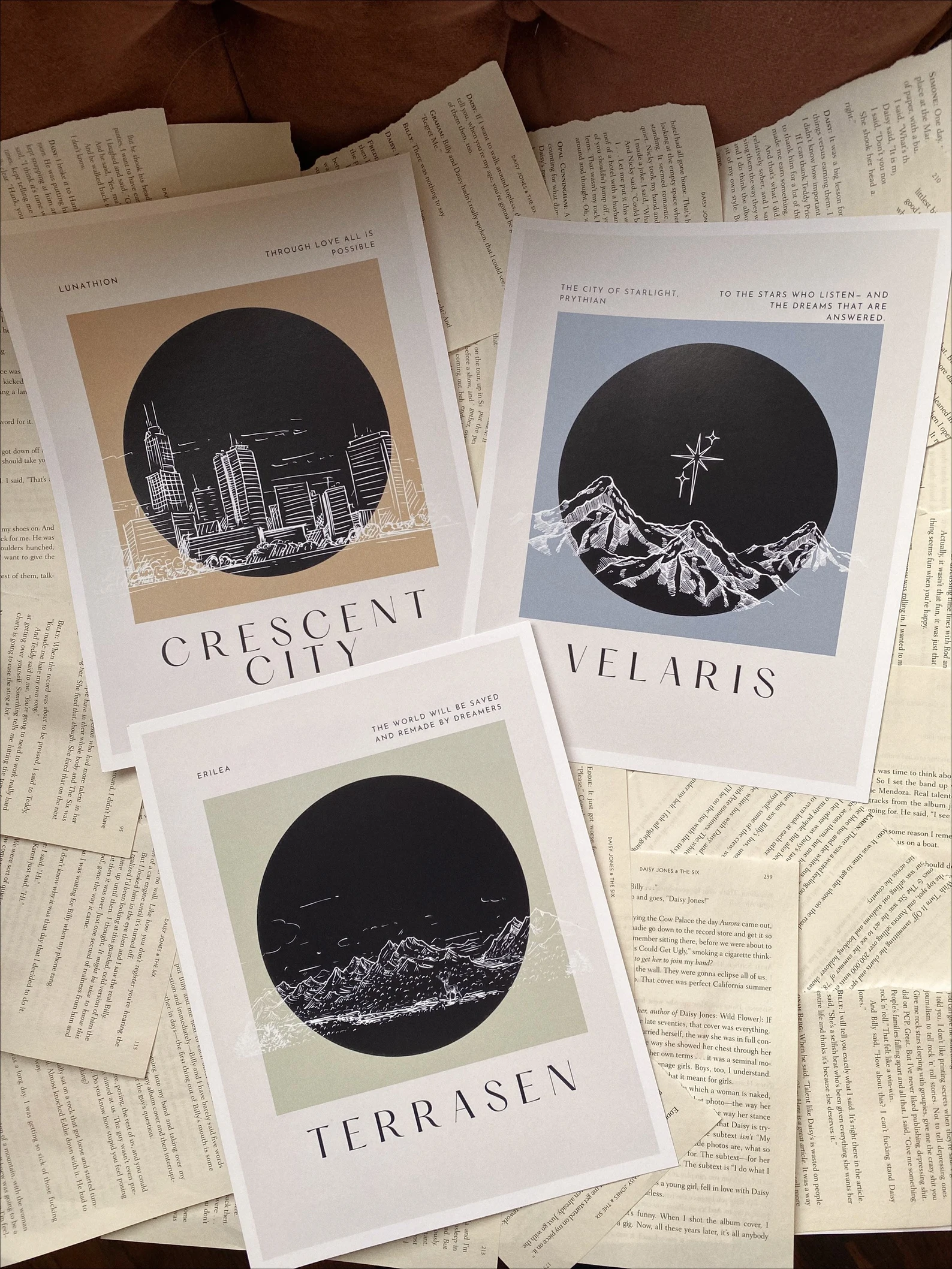 A photo of 3 white postcards. Top to bottom, left to right, the postcards are orange and contain a city scape with the words Crescent City, blue and contain mountains and a star with the words Velaris, and green and contain mountains with the words Terrasen.