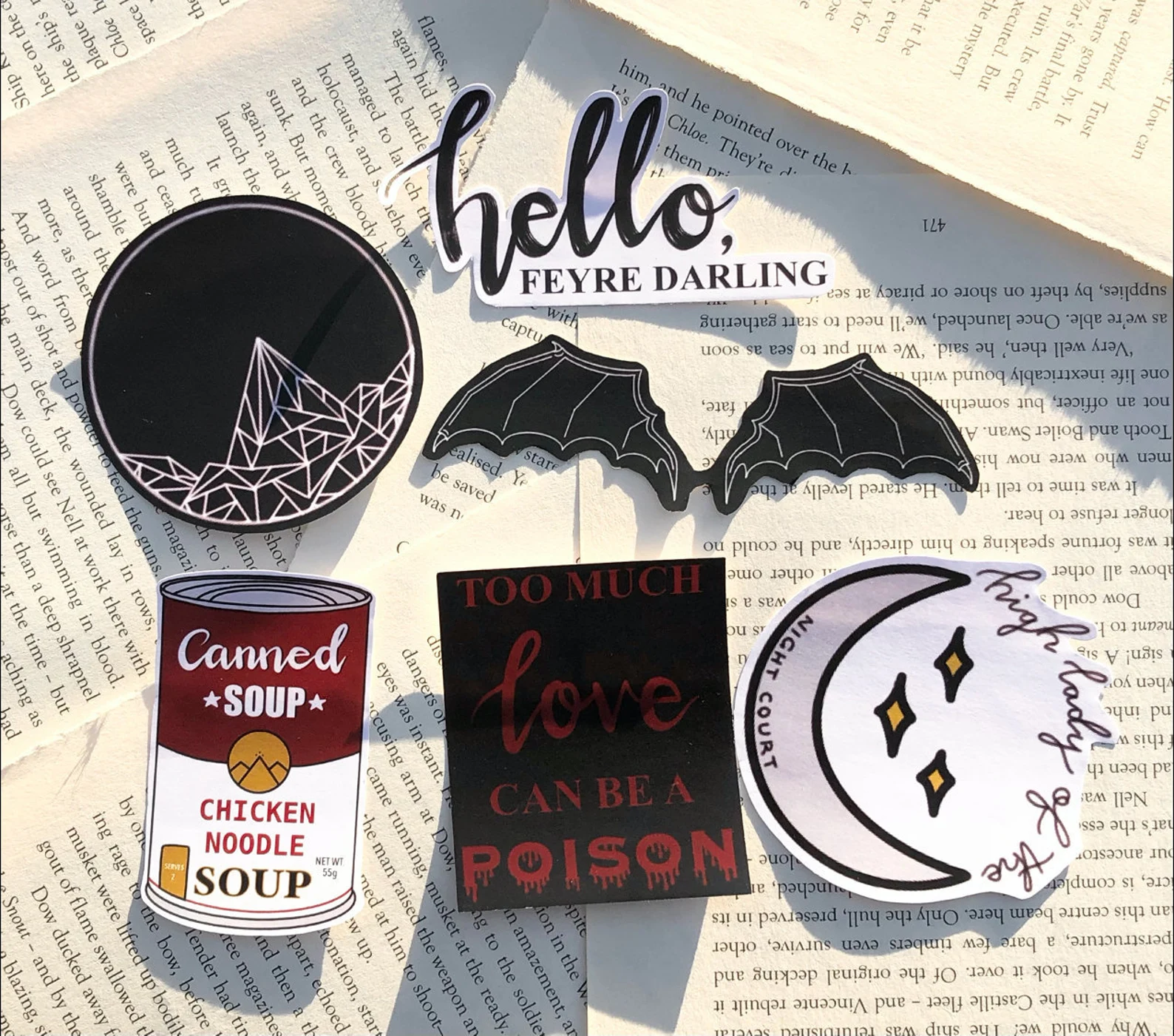A photo of 6 Stamp Stickers ontop of pages. Top to bottom, left to right, the stickers are a round black and white mountian, the words “hello, Feyre darling,” a set of black and white bat wings, a can of soup, the words “too much love can be poision,” and the words high lady of the night court on a moon with stars. 