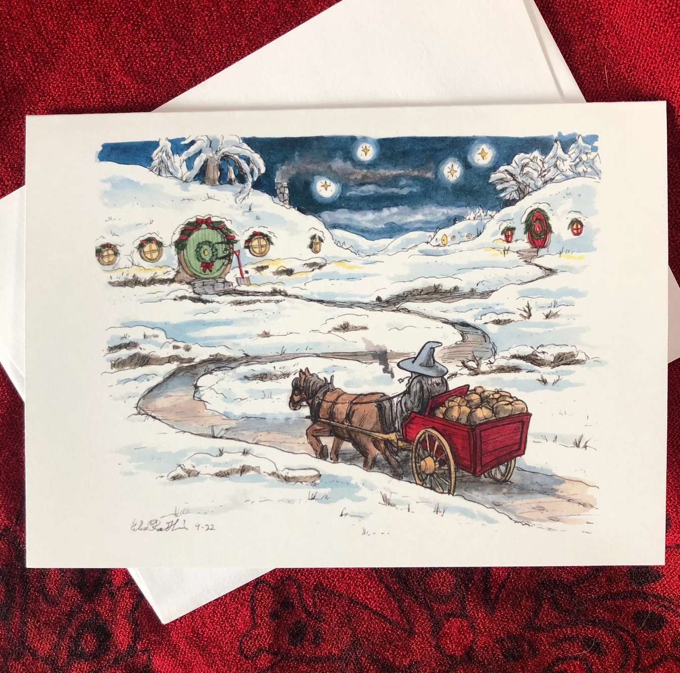 A holiday card featuring a beautiful watercolor and ink drawing of Gandalf showing up in the snow-covered Shire with a cartload of gifts. Hobbit holes have garlands on them, and warm lights from the windows welcome him for the celebration