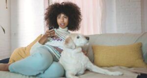 Black woman with full kinky curly afro watches TV on the couch with her dog