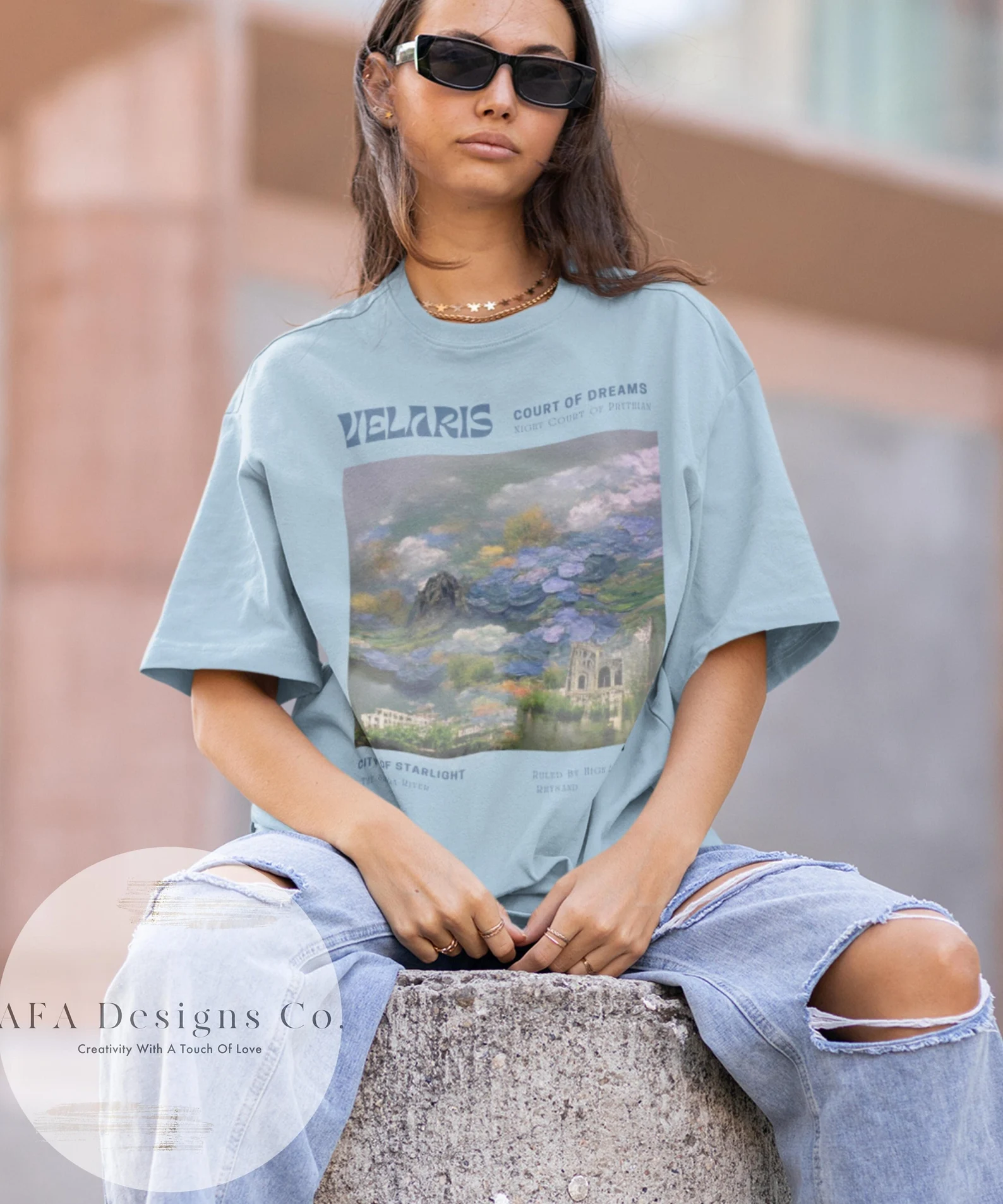 A photo of a person with long brown hair sitting on a pilar wearing a teal shirt with a rectangular image of a pastel landscape and the words Velaris, court of dreams, night court of Prythian at the top of the box and city of starlight at the bottom on the box. 