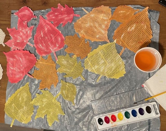 Leaf shapes cut from old books have been waterpainted shades of red, orange, and yellow. They're on a plastic sheet next to the watercolor set and a cup of water.