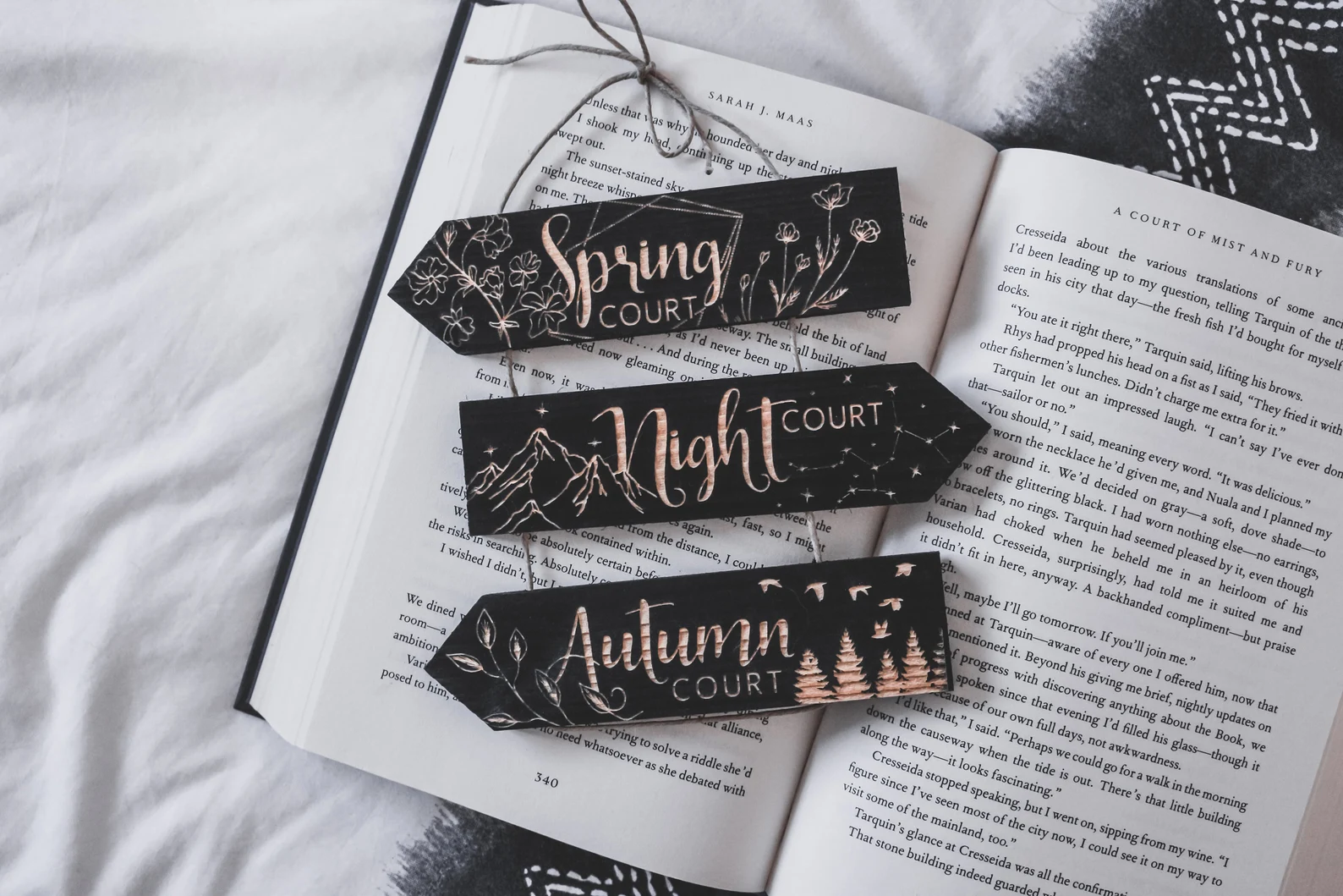 A photo of three black wooden signs stacked and connected with twine. Top to bottom, the signs read Spring Court, Night Court, Autumn Court. The sign is staged on top of a white blanket and an open book. 