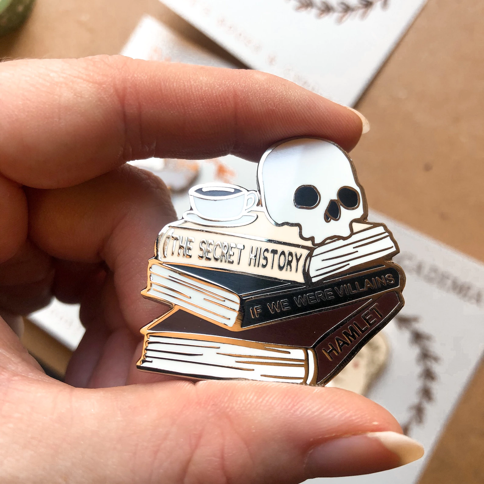 an enamel pin of a stack of books with spines reading "The Secret History," "If We Were Villains," and "Hamlet" with a skull and a cup of tea.