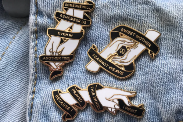 Three enamel pins of gold outlined white hands with black banners wrapping around them featuring various quotes from the poet Sappho. 