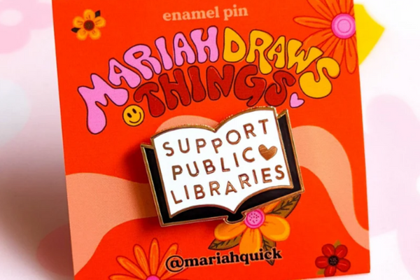 A white enamel pin in the shape of an open book with a black outline and the text "support public libraries" alongside a little heart in gold.