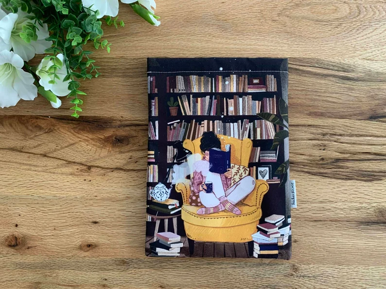 Photo of a black book sleeve with a print of a girl sitting on a yellow chair reading a book, a shelf filled with books behind her