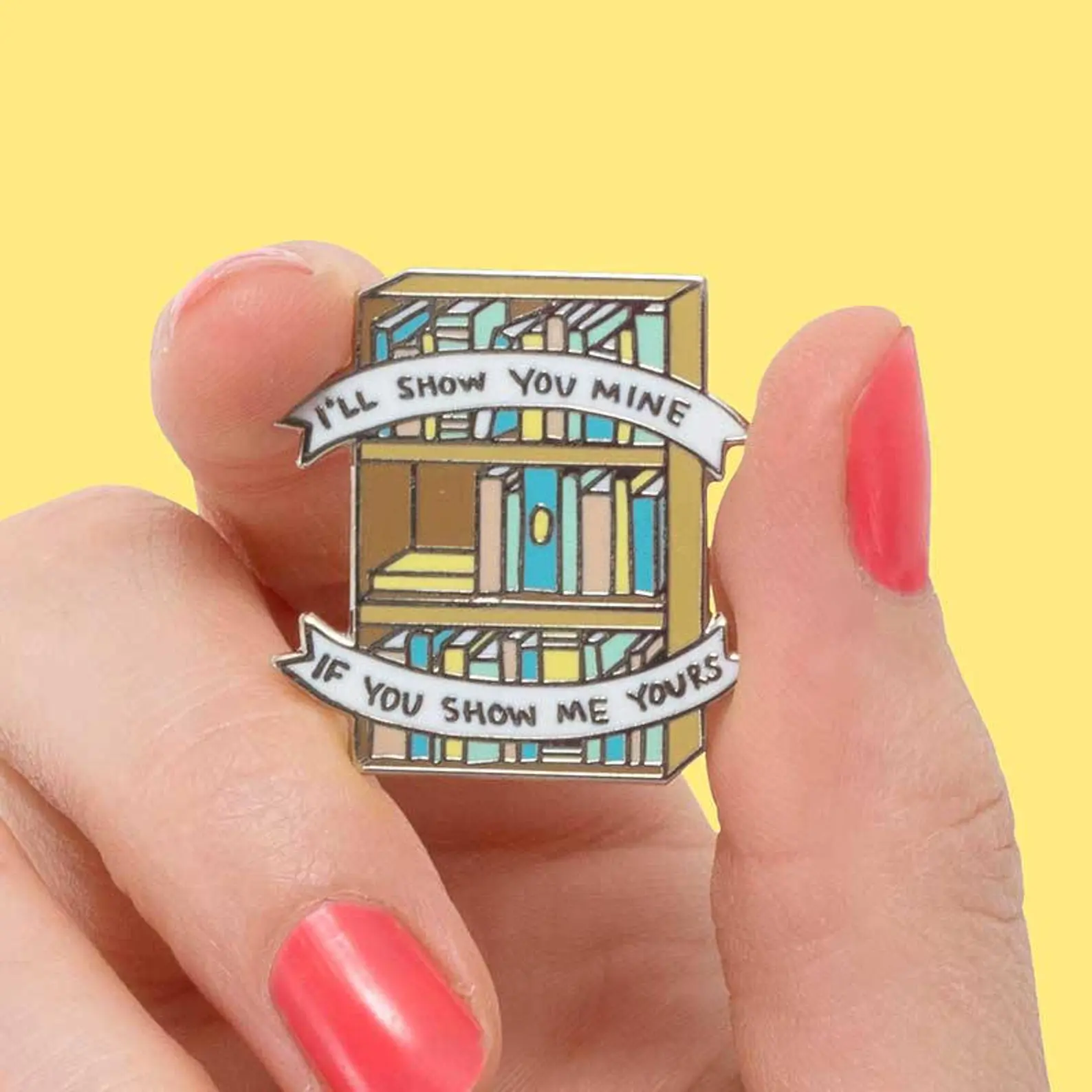 an enamel pin of a bookshelf and a banner that reads "I'll show you mine if you show me yours"