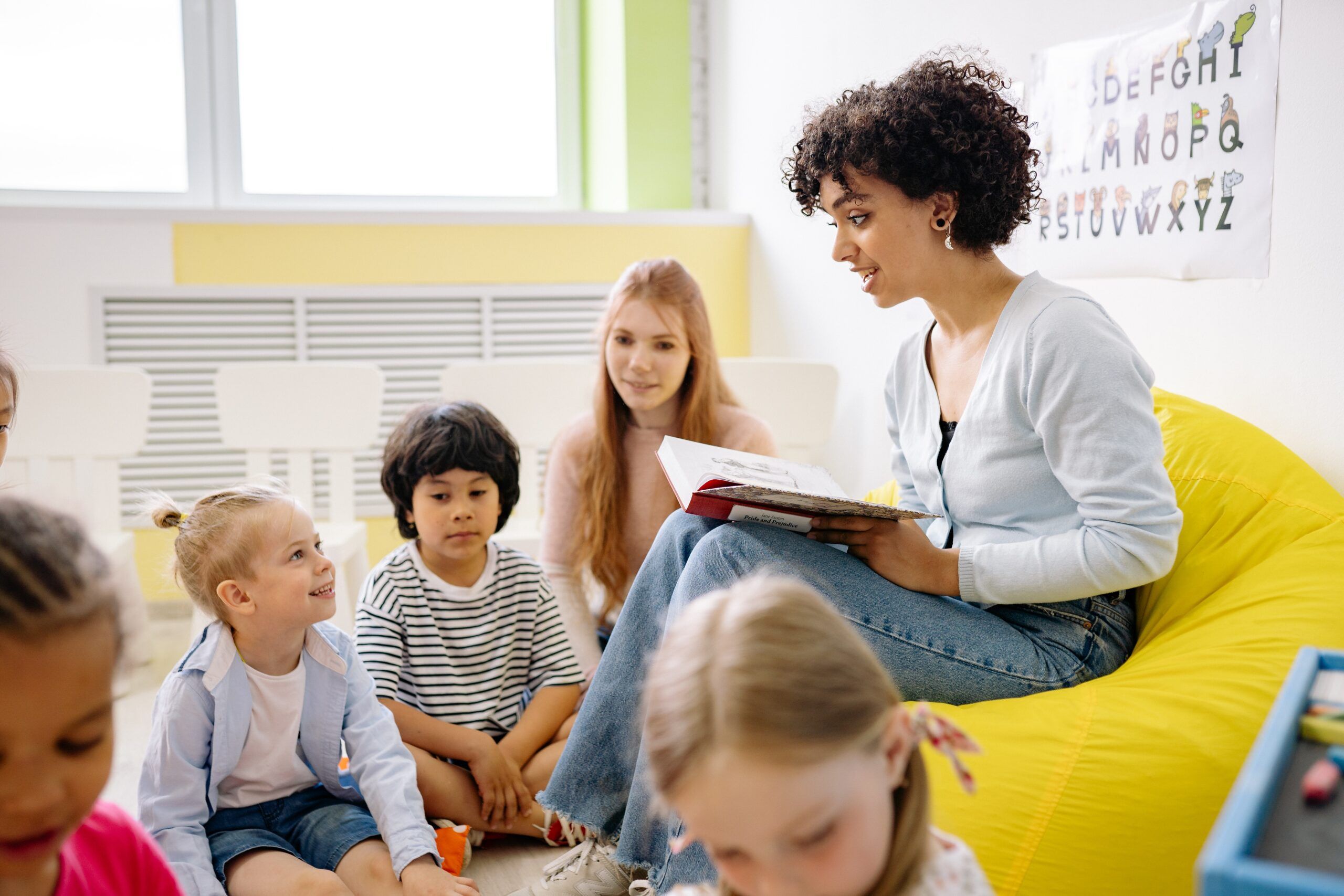 woman of color with kinky curly hair sits on bean bag reading to small children