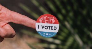 a finger with a red, white, and blue circular sticker with the words "I voted" in the center