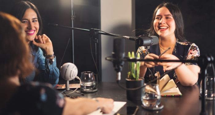 light tan-skinned women with dark hair are smiling and sitting at a table with microphones and books