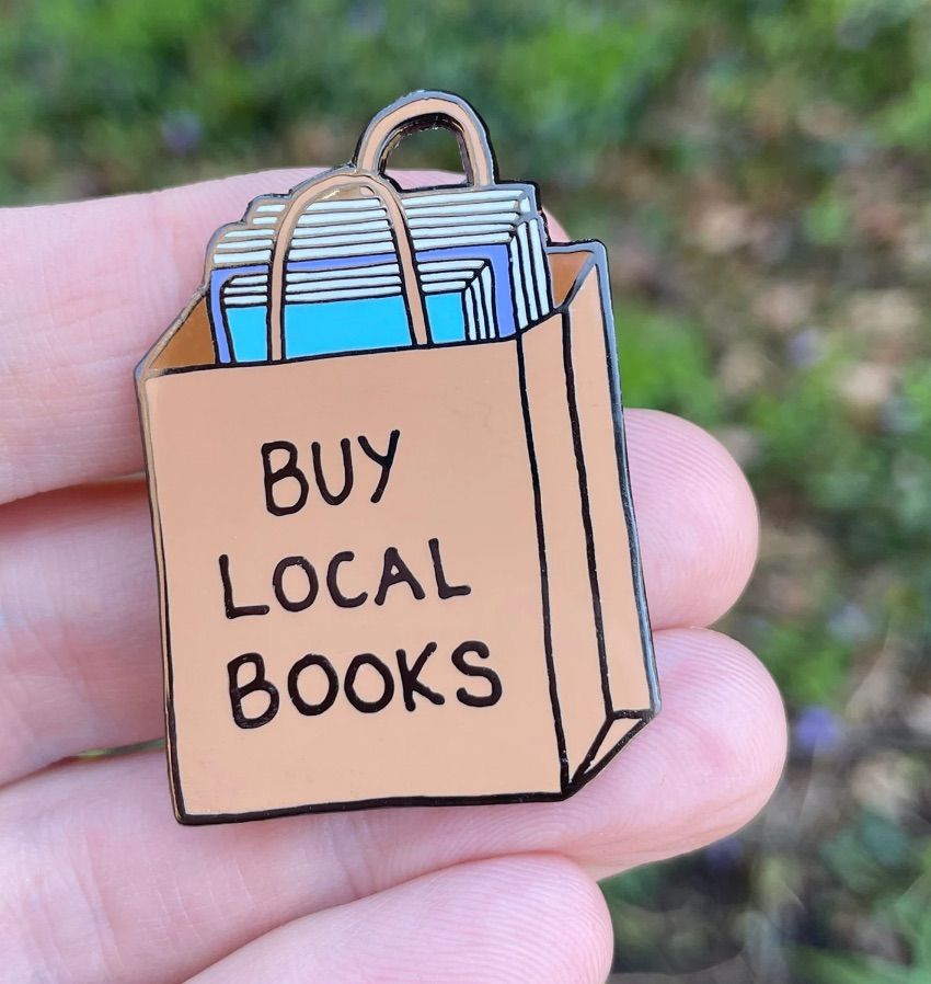 Image of an enamel pin in the shape of a brown paper bag. There are books peeking out from the top. The bag says "buy local books."