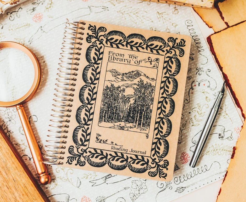 Image of a notebook that looks like an old book. It is a custom reading journal, sitting on a table beside a pen and magnifying glass. 