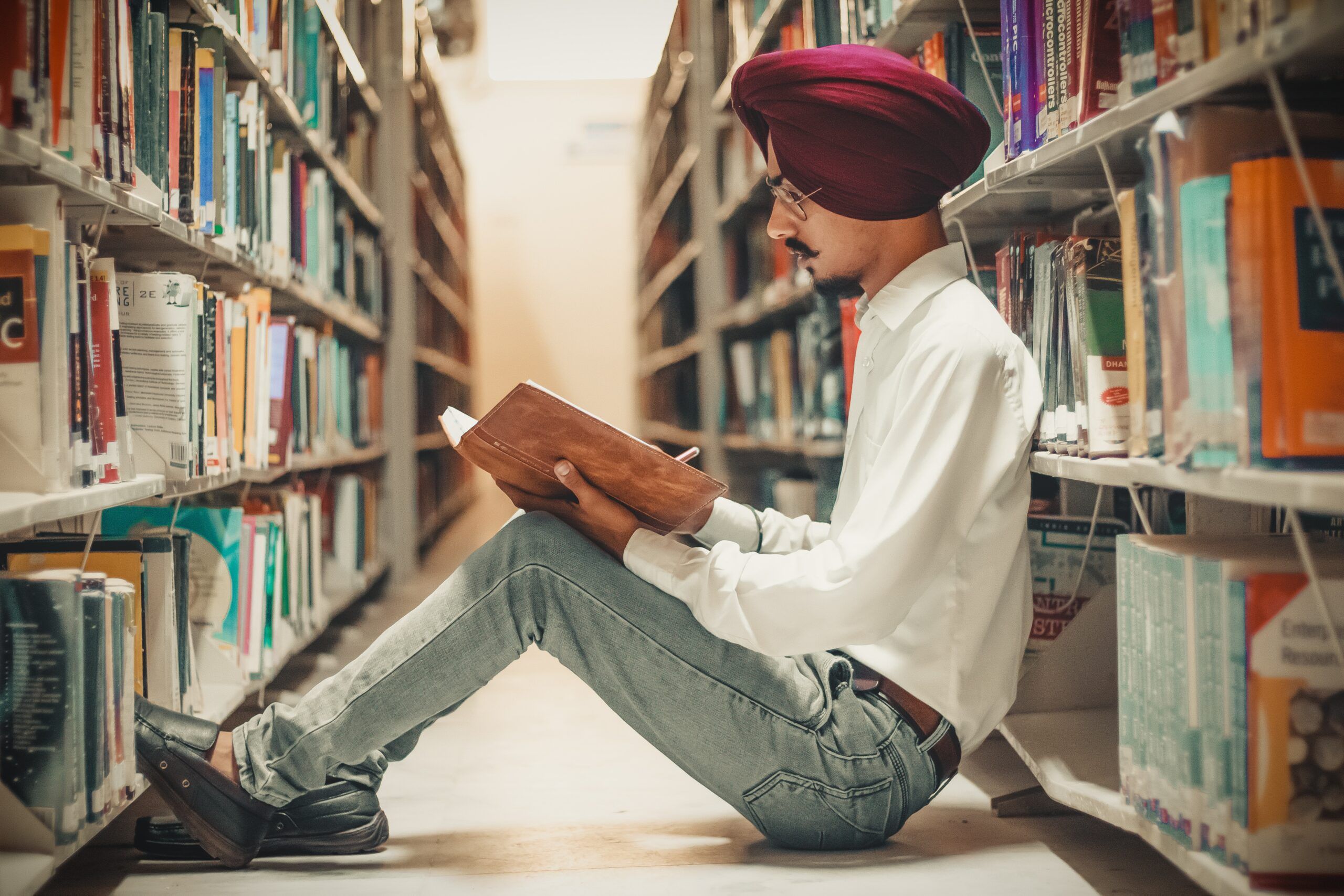 Tan-skinned man with a burgundy turban is sitting in a library reading