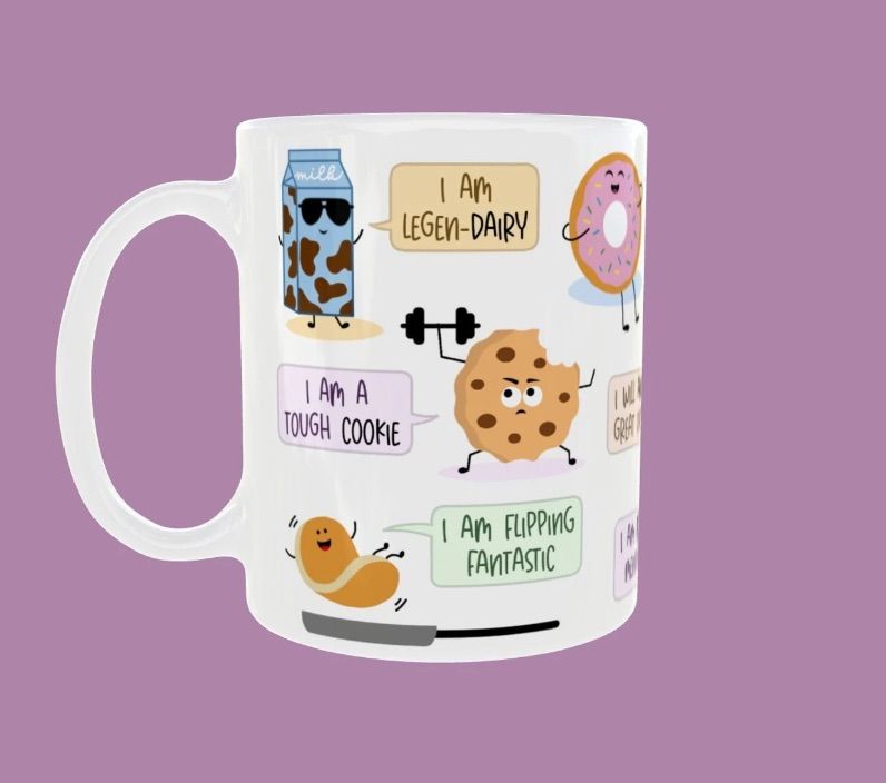 White mug on a purple background. The mug features several foods, including cookies, pancakes, and milk, with assorted affirmation puns related to them, such as "I am a tough cookie."