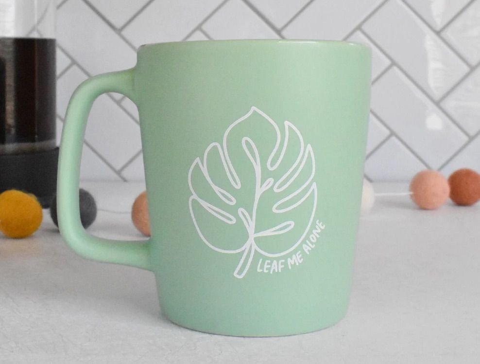 Image of a green mug on a white counter. In white, there is an image of a plant leaf with the text "leaf me alone."