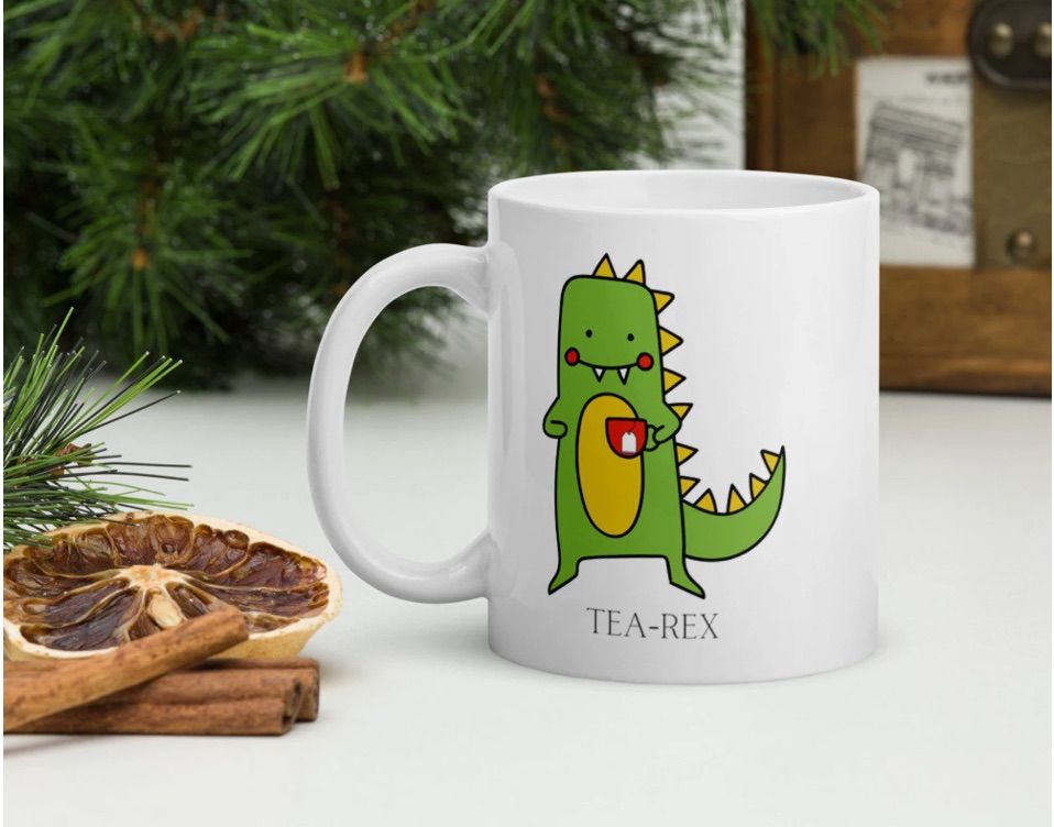 Image of a white mug in front of garland. It has a green t-rex on it, with a red mug of tea. Beneath the dino it reads "tea-rex."