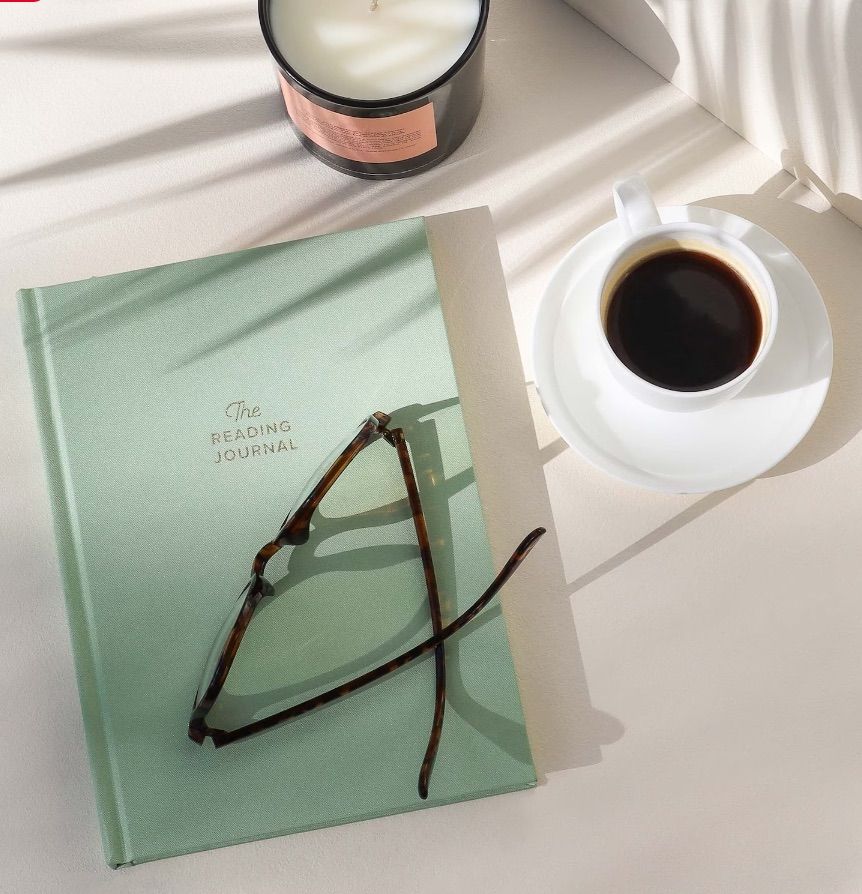 Image of a green reading journal. It has a pair of glasses on top of it, with a coffee cup beside it. 