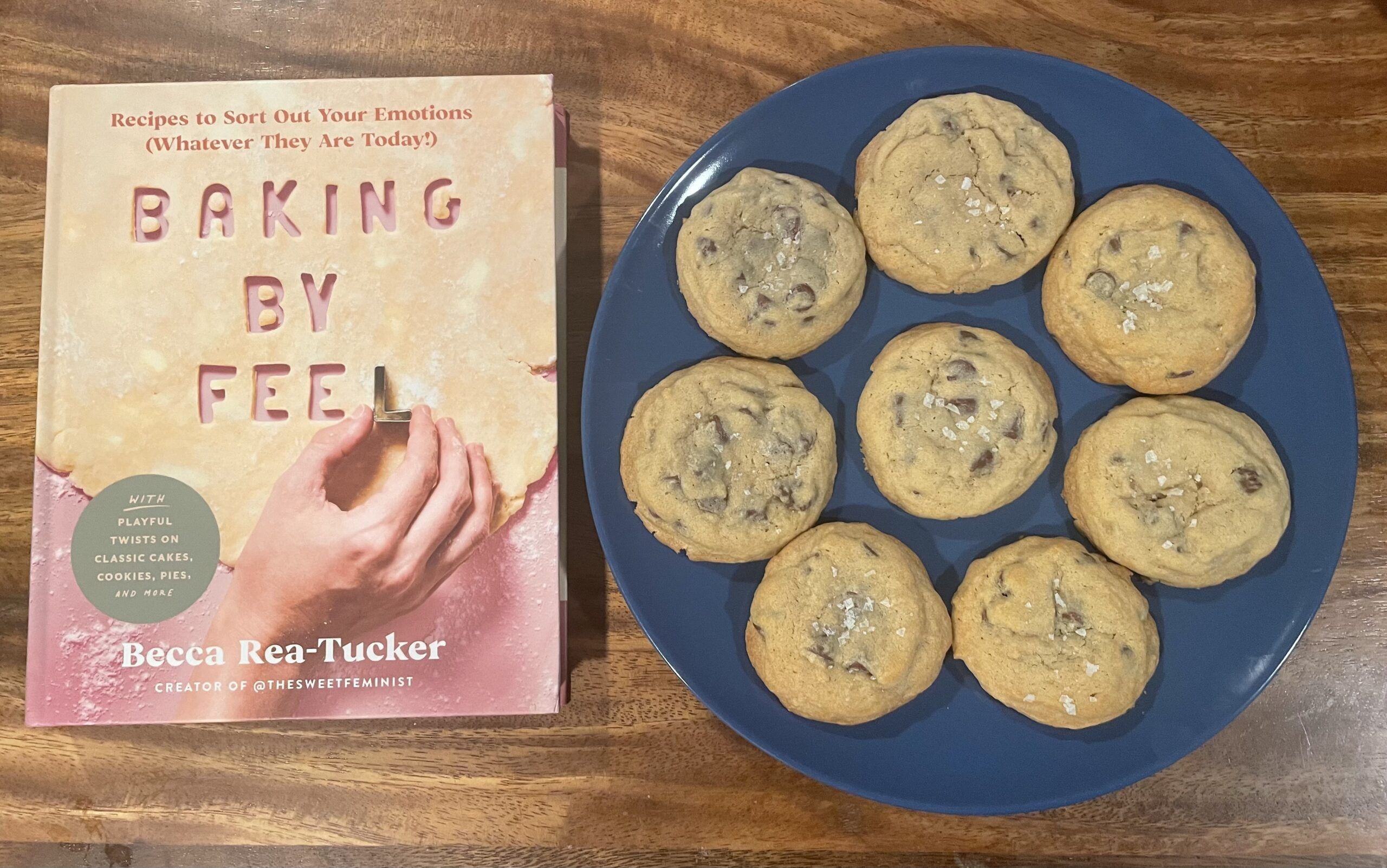 Baking By Feel cookbook on a wooden table next to a plate of fluffy, light brown chocolate chip cookies