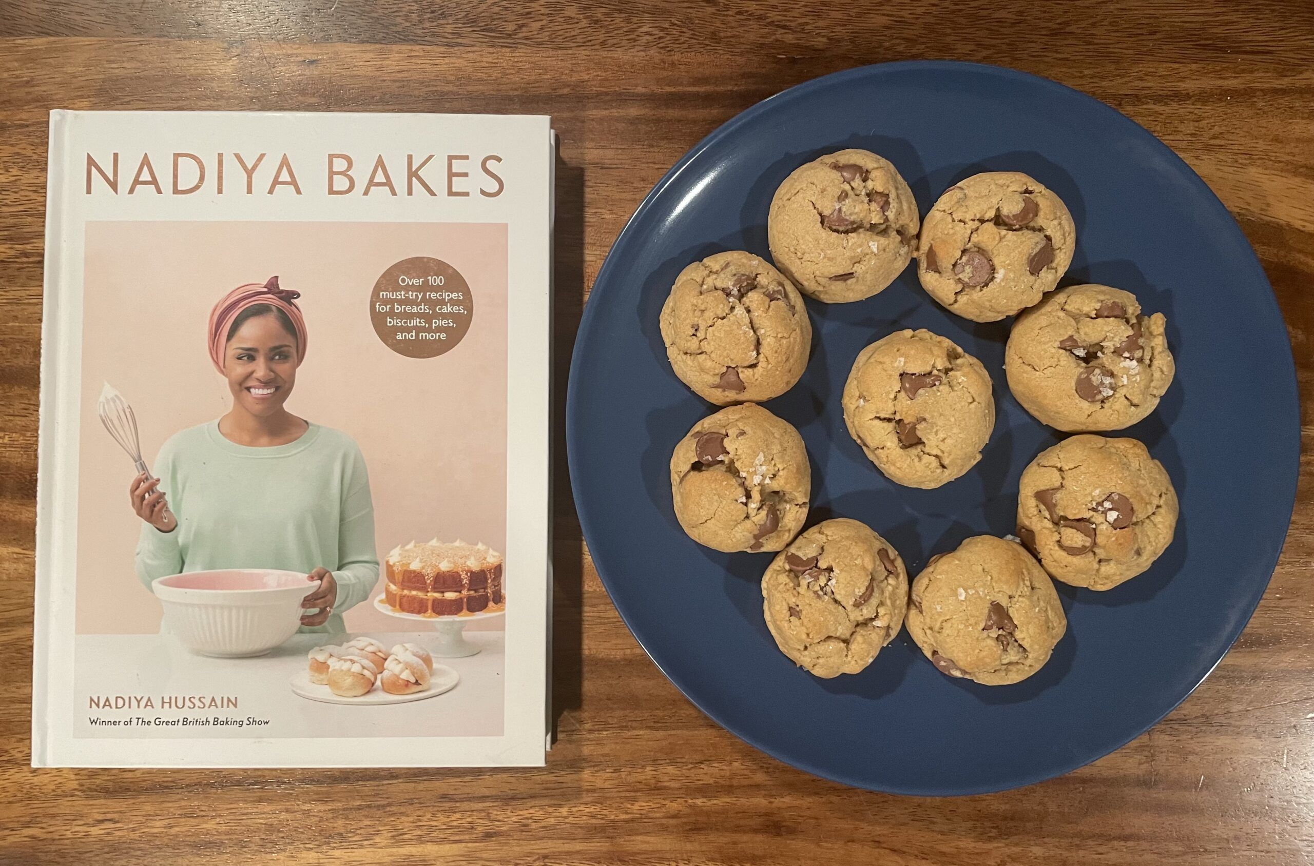 The cookbook Nadiya Bakes is on a wooden table next to a blue plate of small, rounded chocolate chip cookies.