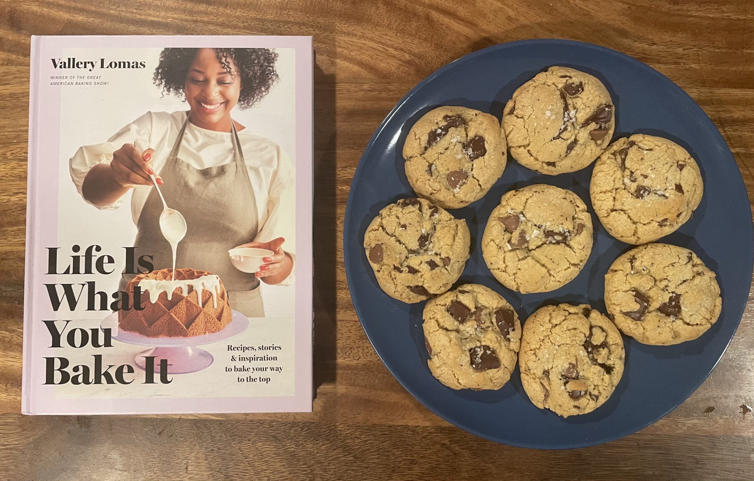 Life is What You Bake It cookbook on a wooden table next to a plate of chocolate chunk cookies.