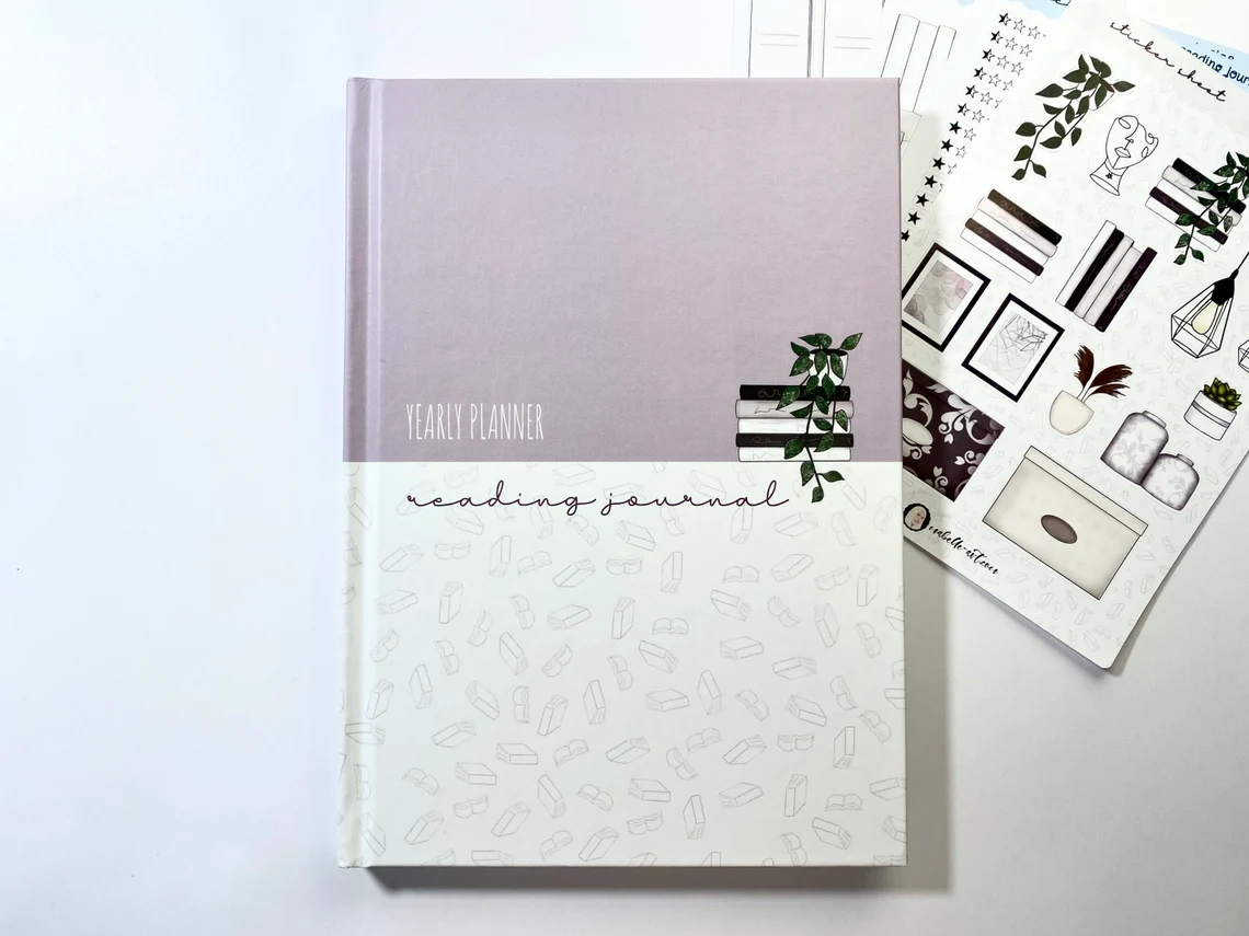 Picture of a pink and white journal with an illustration of a stack of books and a plant on top. Text: Yearly planner, reading journal