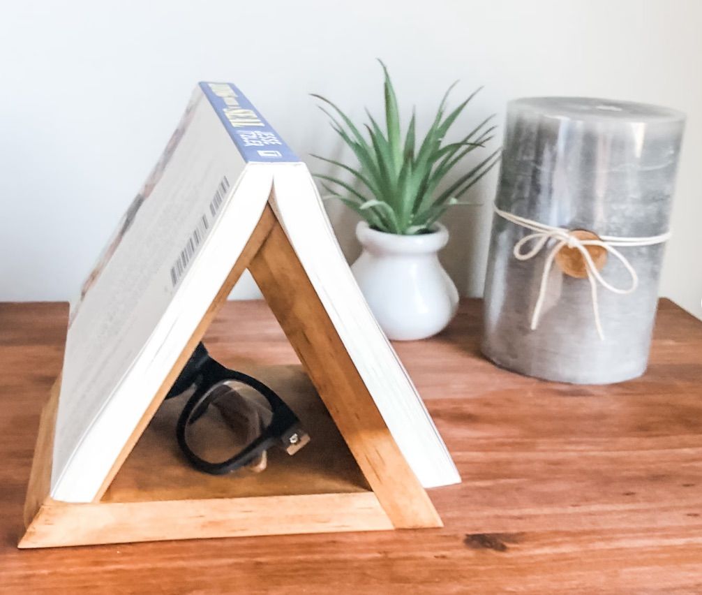 Image of a nightstand. On it is a wooden triangle that allows a book to rest open on top, with space for glasses beneath. 