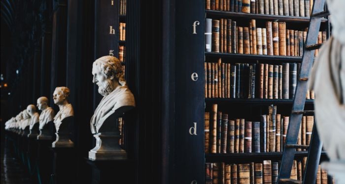 row of dark wooden shelves filled with books in a library with marble busts at the entrance to each row