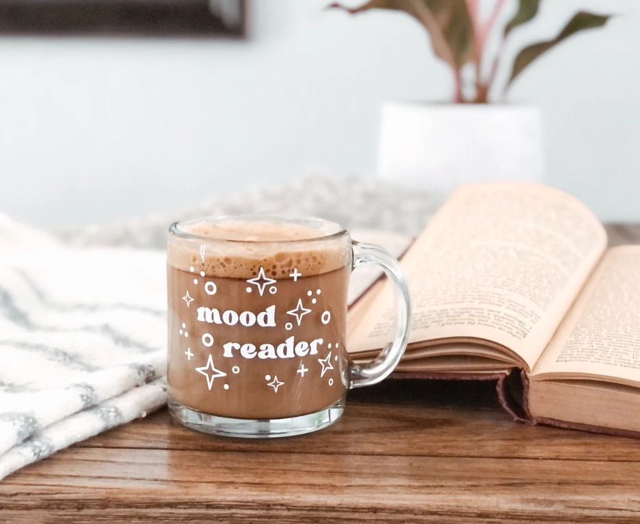 Image of a glass mug on a table with an open book. In white on the mug are the words "mood reader."